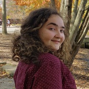 A headshot of a female team member. Her body is titled to the side, with her head angled towards the camera. She has curly dark brown hair, pale skin, and is smiling at the camera.
