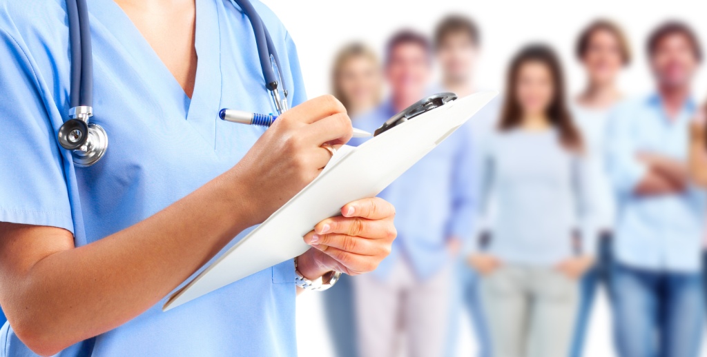 A cropped image a medical professional in scrubs writing on a cripboard with a blue pen. They have tanned skin, are wearing a silver watch, and have a stethoscope around their neck. In the background, there is a group of blurred people wearing casual clothes, standing in uniform lines looking relaxed.