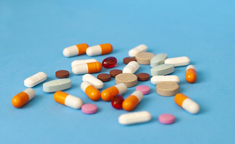A pile of loose pills laying on a blue background. There are a variety of pill colors and shapes, consisting of red, orange, white, tan, and pink long and flat pills.